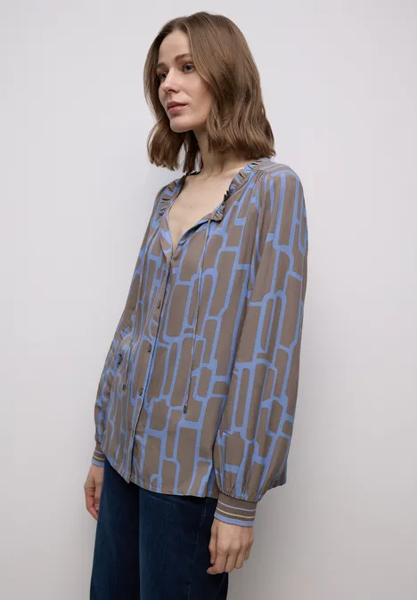 Frau in geometrisch gemusterter, braun-blauer Bluse und Jeans vor weißem Hintergrund.